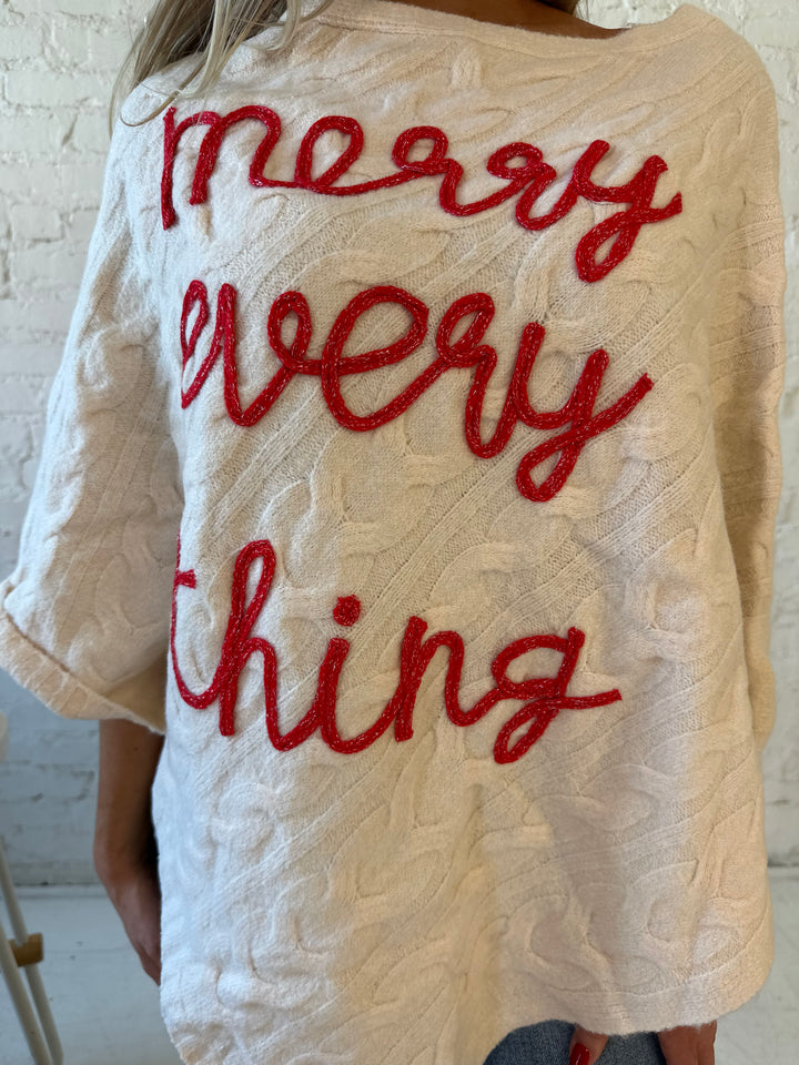 Woman wearing an ivory cable knit poncho with the words merry everything in red cursive. 