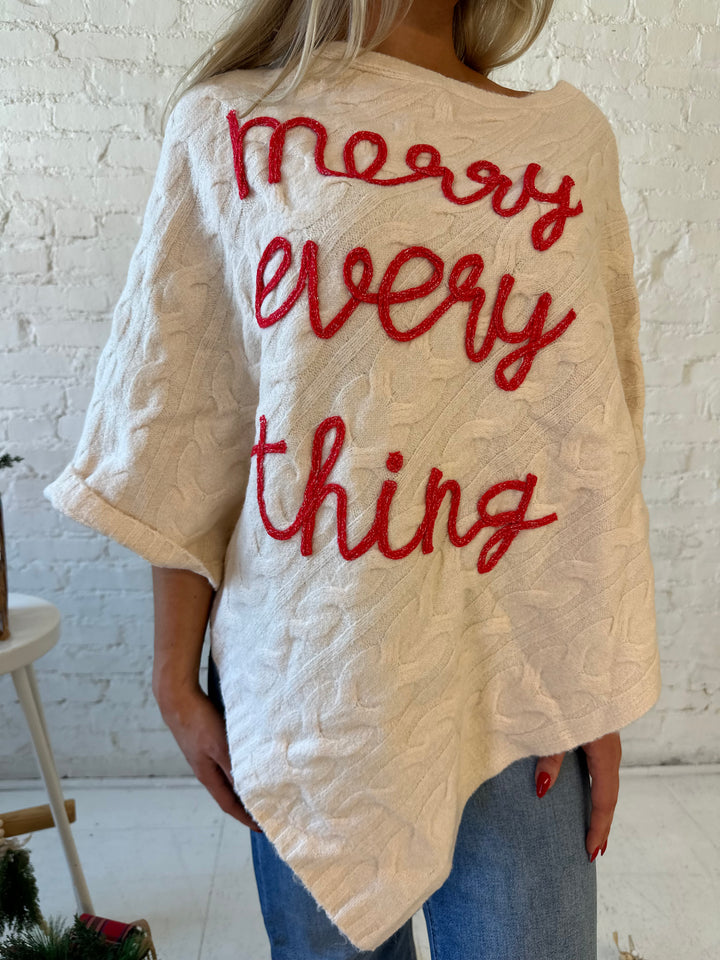 Woman wearing an ivory cable knit poncho with the words merry everything in red cursive. 
