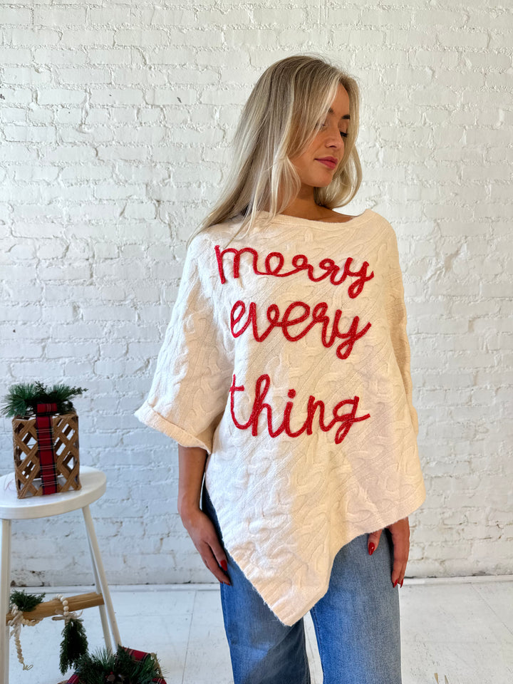 Woman wearing an ivory cable knit poncho with the words merry everything in red cursive. 