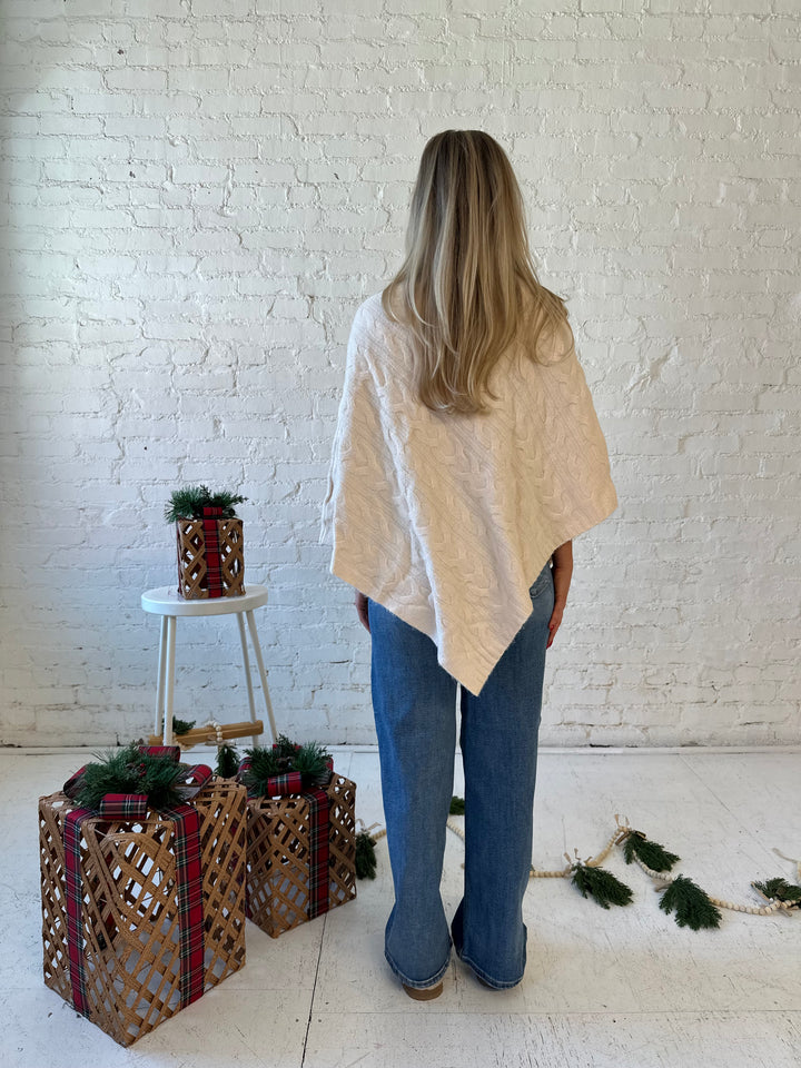 Woman wearing an ivory cable knit poncho with the words merry everything in red cursive. 