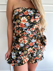 Woman wearing a black floral strapless romper with wrap skirt detail.