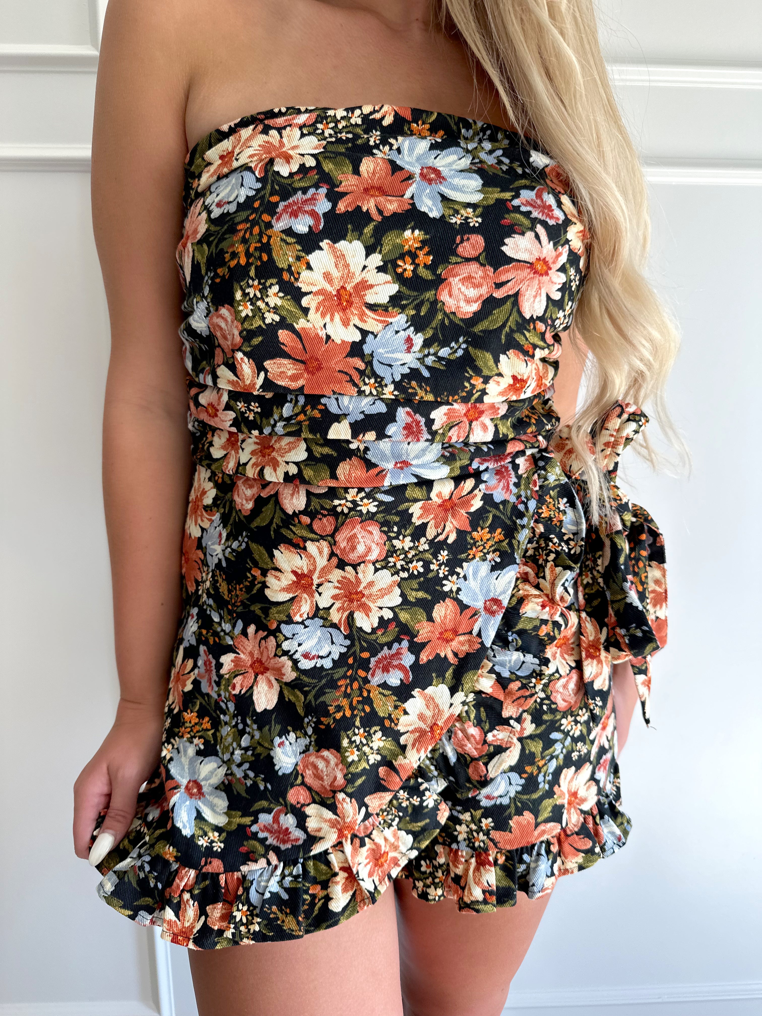 Woman wearing a black floral strapless romper with wrap skirt detail.