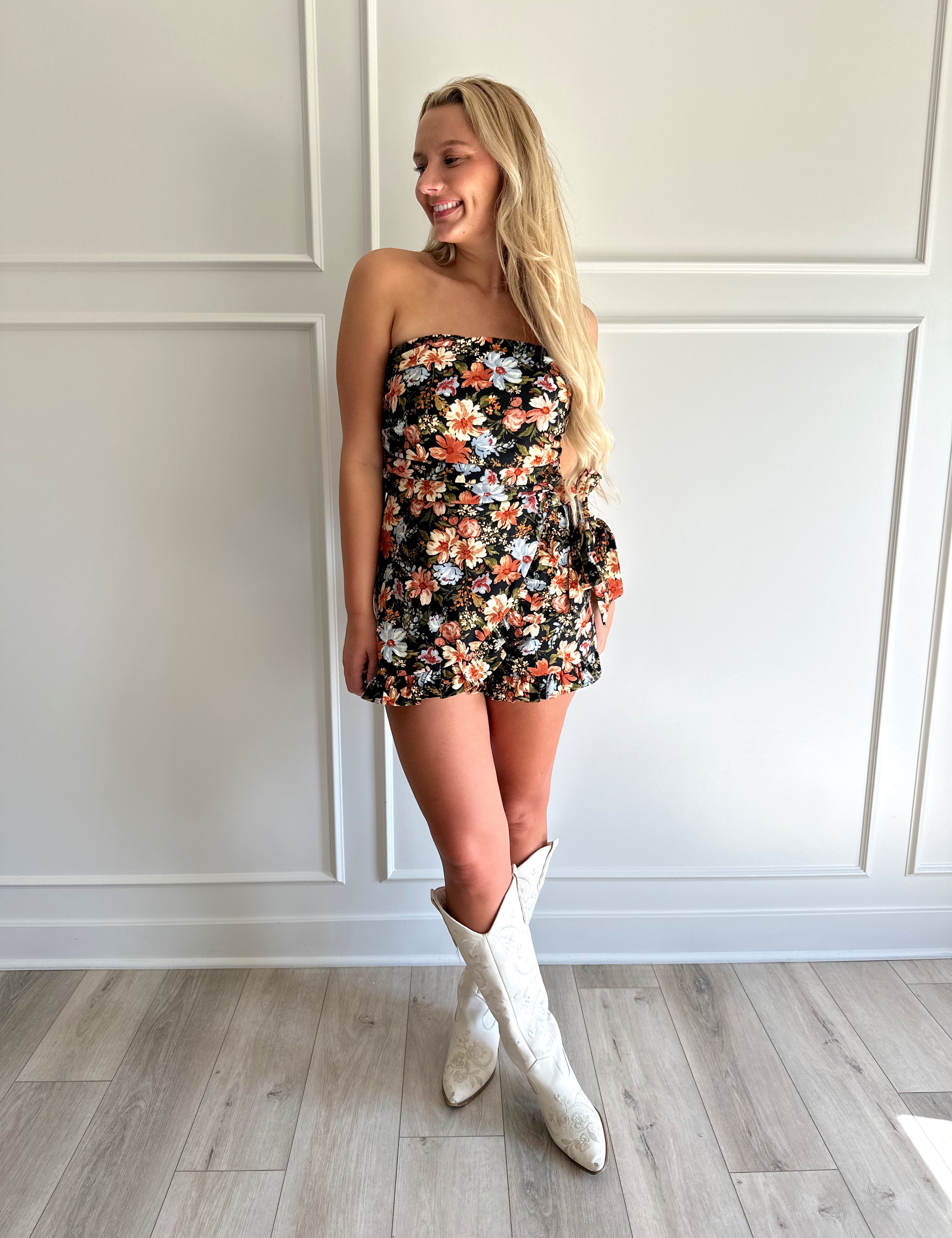 Woman wearing a black floral strapless romper with wrap skirt detail.