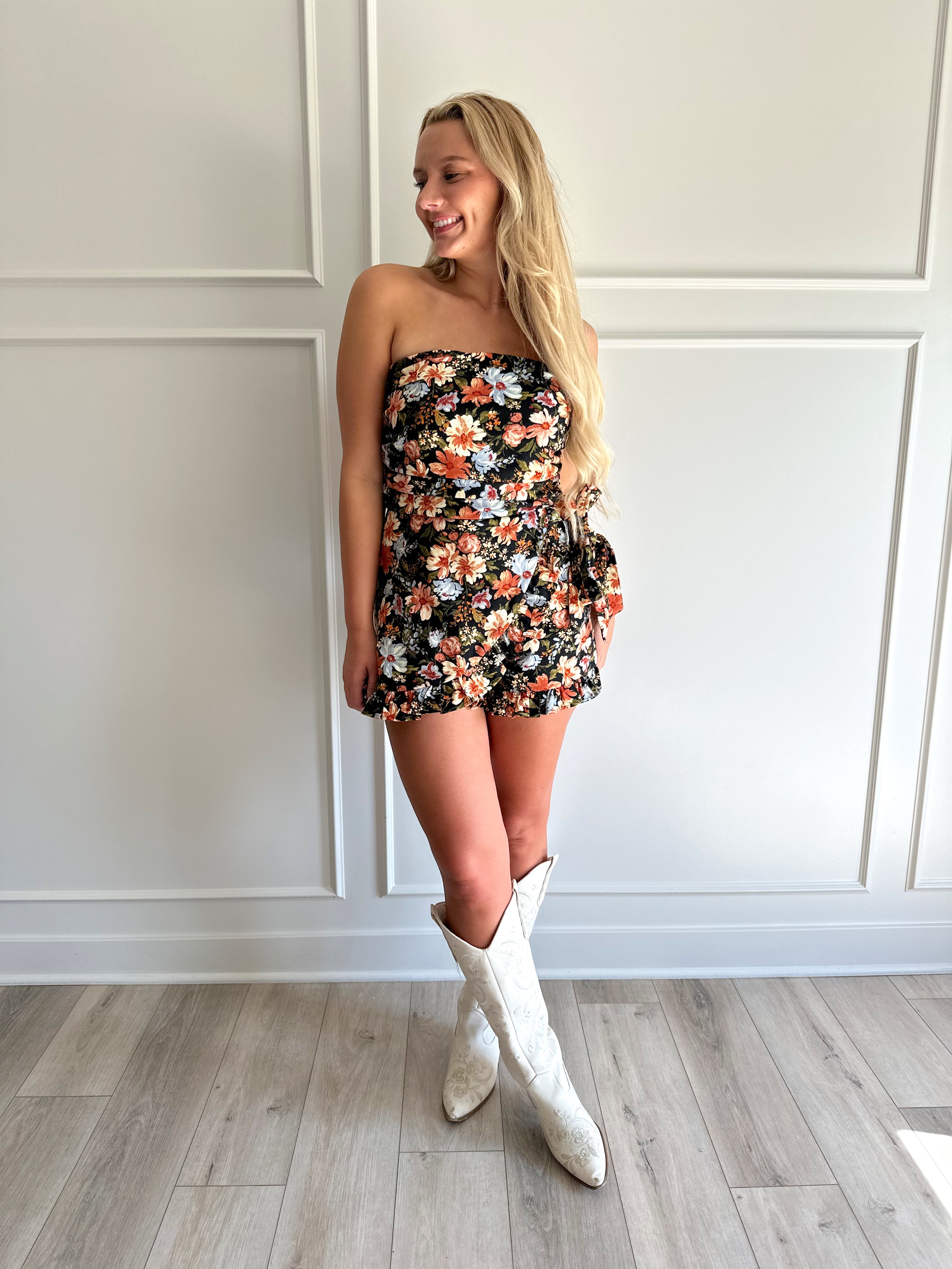 Woman wearing a black floral strapless romper with wrap skirt detail.
