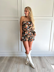Woman wearing a black floral strapless romper with wrap skirt detail.
