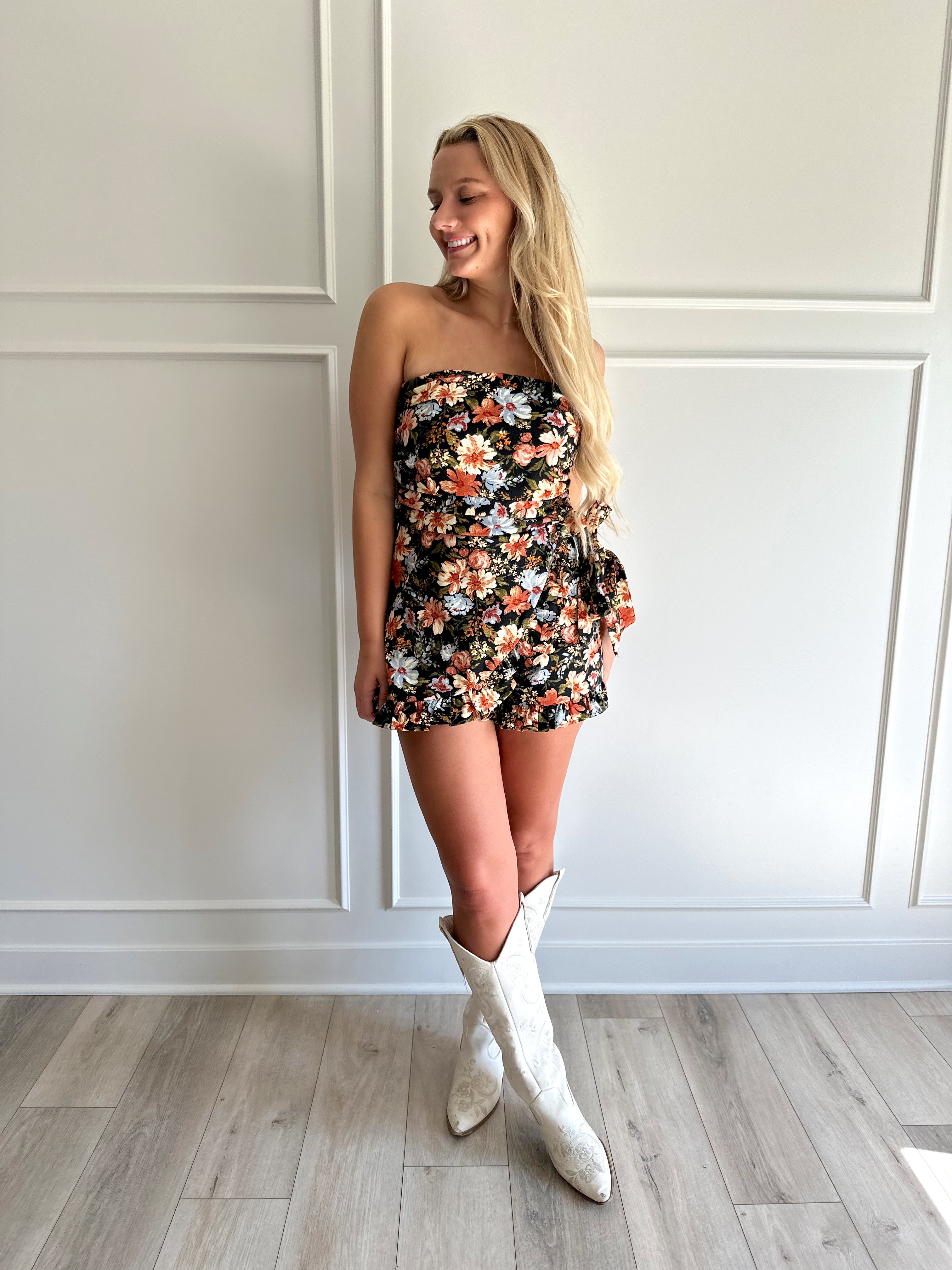 Woman wearing a black floral strapless romper with wrap skirt detail.
