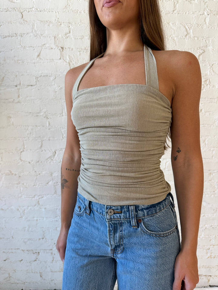 Woman wearing a taupe ruched halter top with medium wash jeans. 