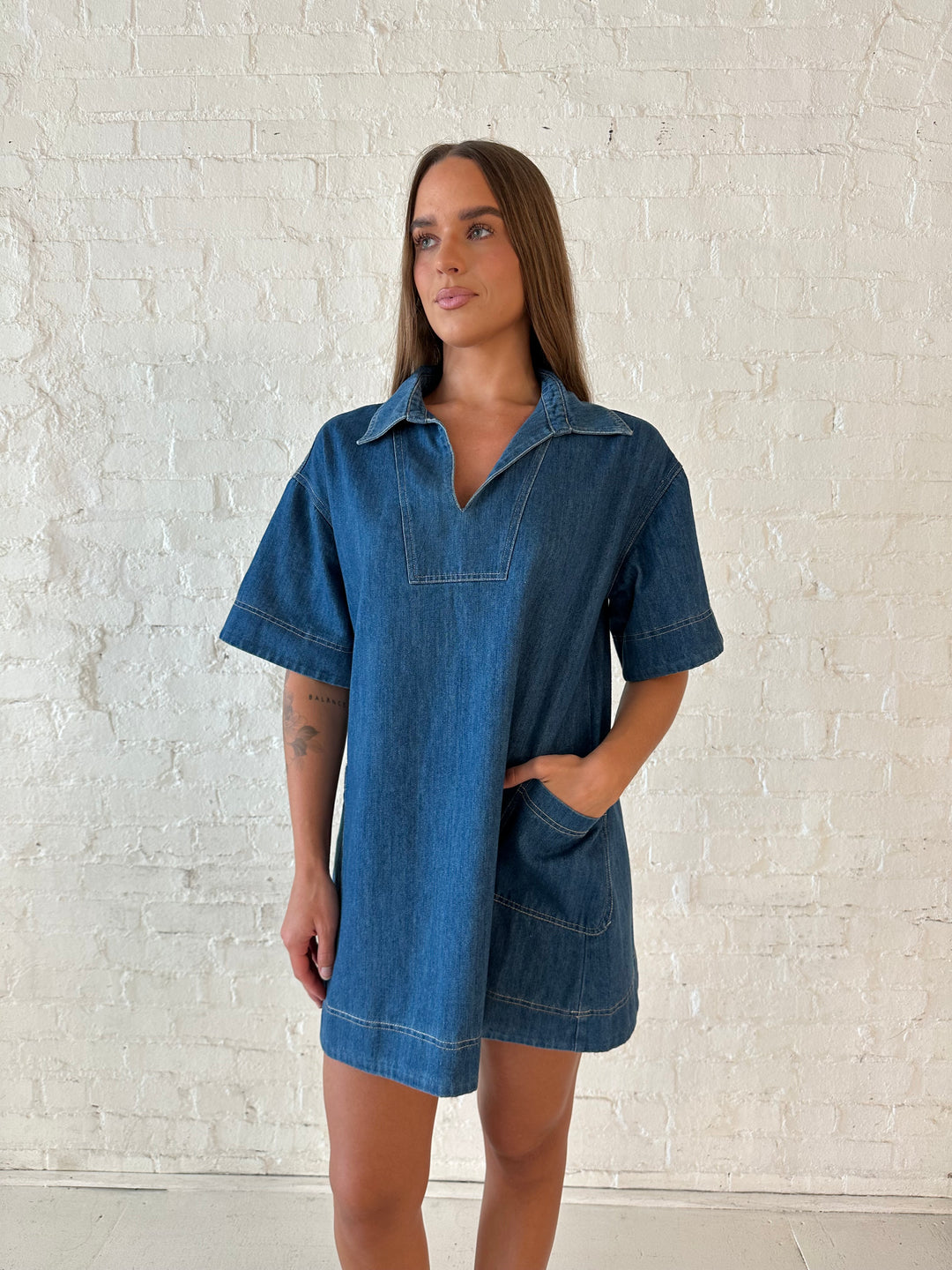 Woman wearing dark wash shift style mini dress with short sleeves, collar detailing, and pockets. 