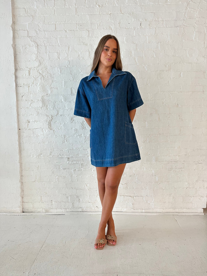 Woman wearing dark wash shift style mini dress with short sleeves, collar detailing, and pockets. 