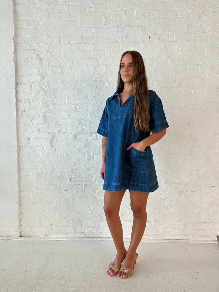 Woman wearing dark wash shift style mini dress with short sleeves, collar detailing, and pockets. 