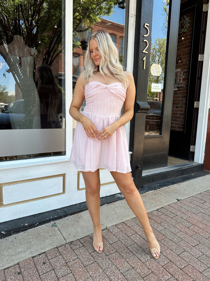 Woman wearing light pink striped strapless mini dress with open back. 