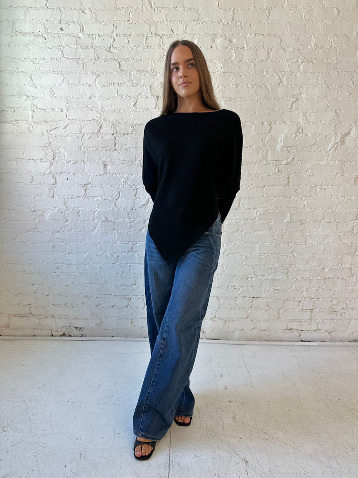 Woman wearing black ribbed long sleeve sweater with pointed hem, and dark wash jeans. 