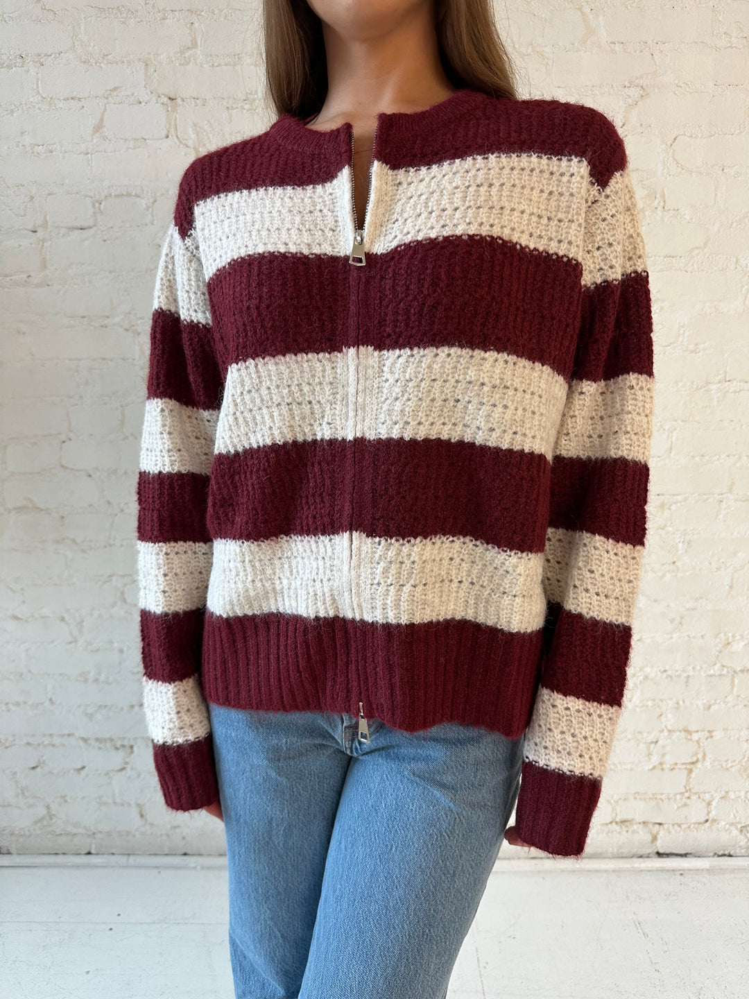 Woman wearing ivory and burgundy chunky striped zip up jacket with medium wash jeans. 