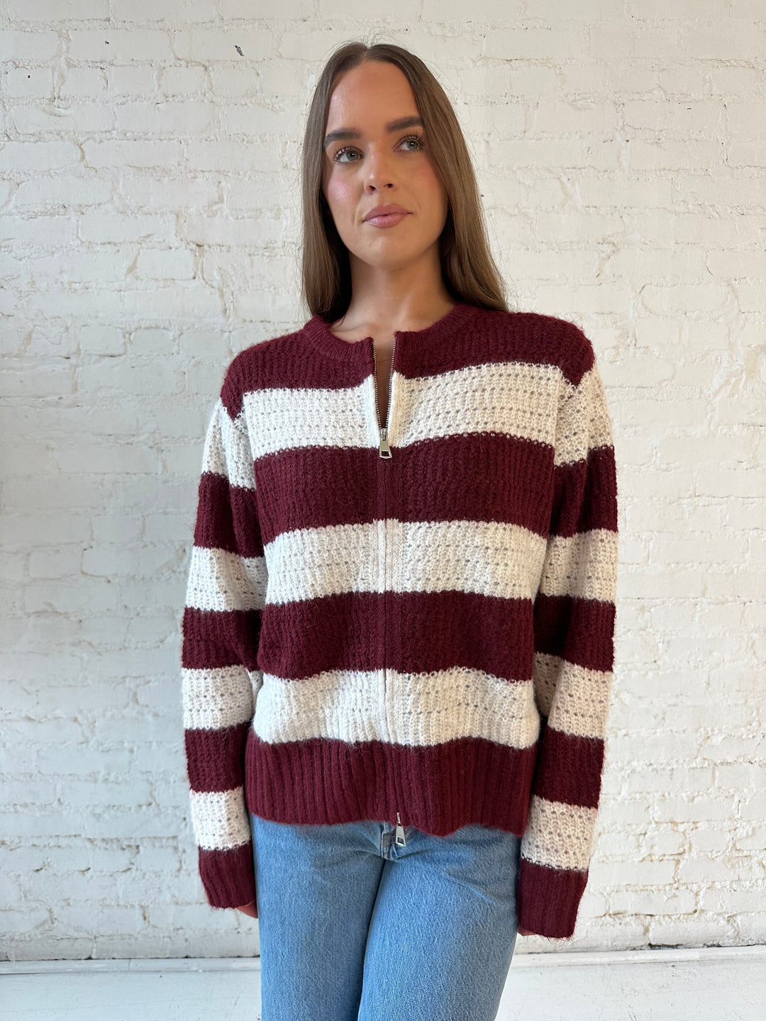 Woman wearing ivory and burgundy chunky striped zip up jacket with medium wash jeans. 