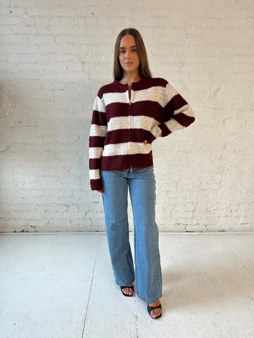Woman wearing ivory and burgundy chunky striped zip up jacket with medium wash jeans. 