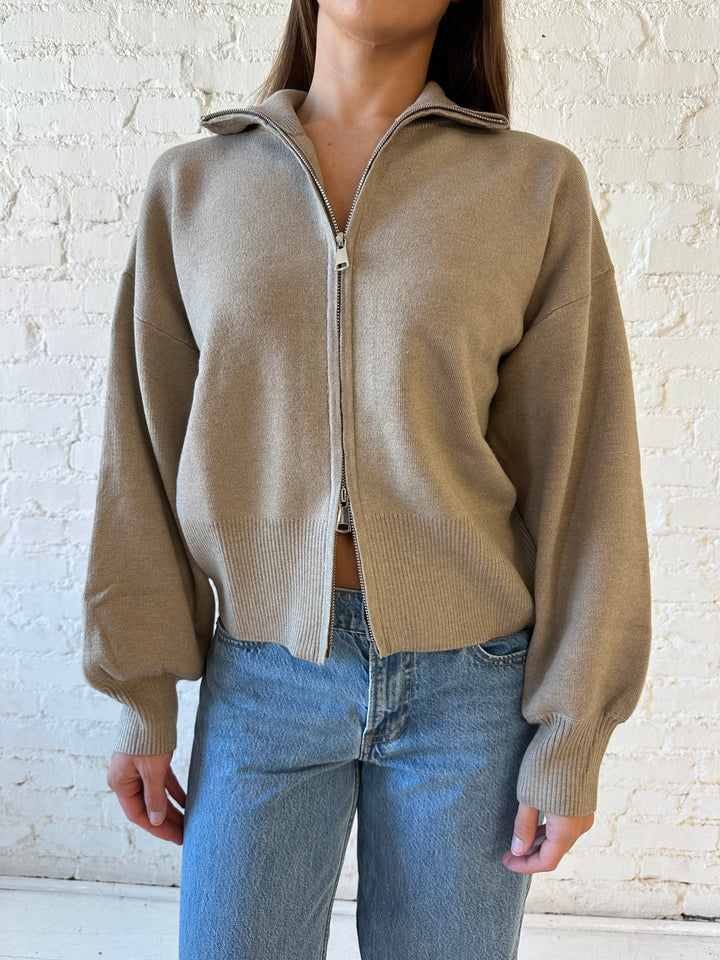 Woman wearing khaki colored knit zip up jacket with medium wash jeans. 