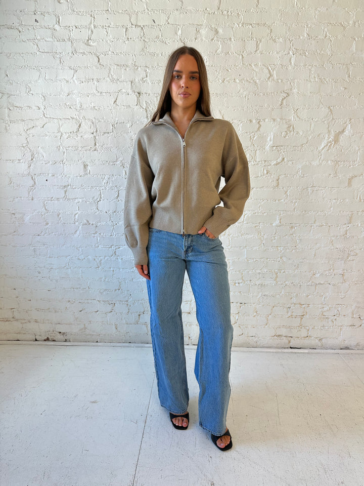 Woman wearing khaki colored knit zip up jacket with medium wash jeans. 