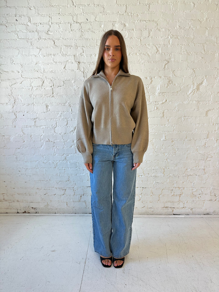 Woman wearing khaki colored knit zip up jacket with medium wash jeans. 