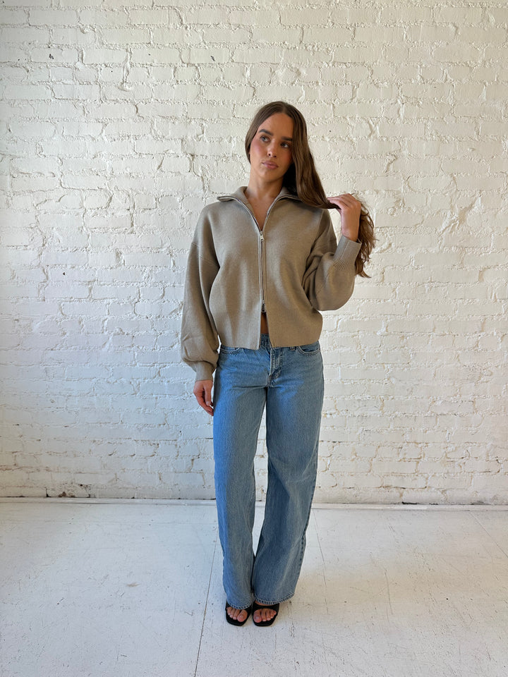 Woman wearing khaki colored knit zip up jacket with medium wash jeans. 