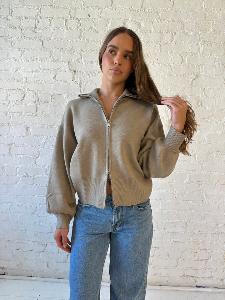 Woman wearing khaki colored knit zip up jacket with medium wash jeans. 