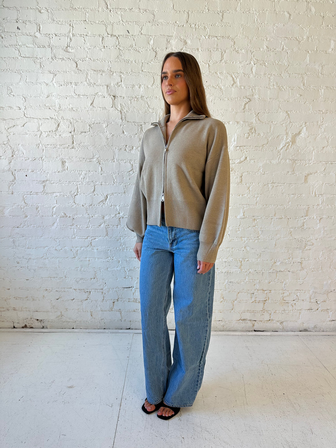 Woman wearing khaki colored knit zip up jacket with medium wash jeans. 
