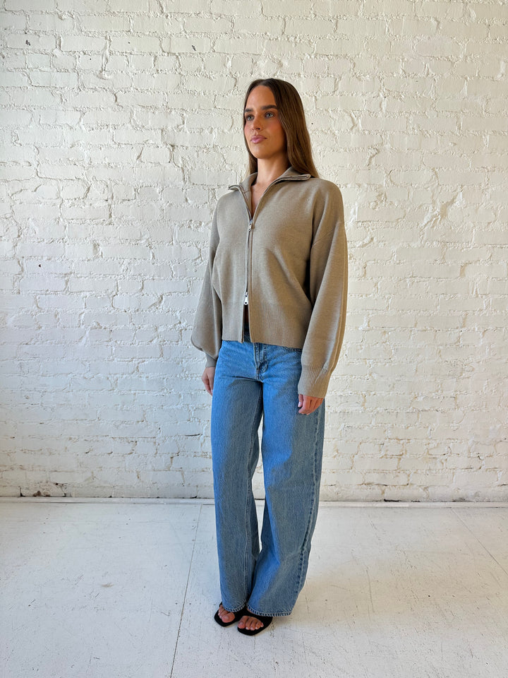 Woman wearing khaki colored knit zip up jacket with medium wash jeans. 