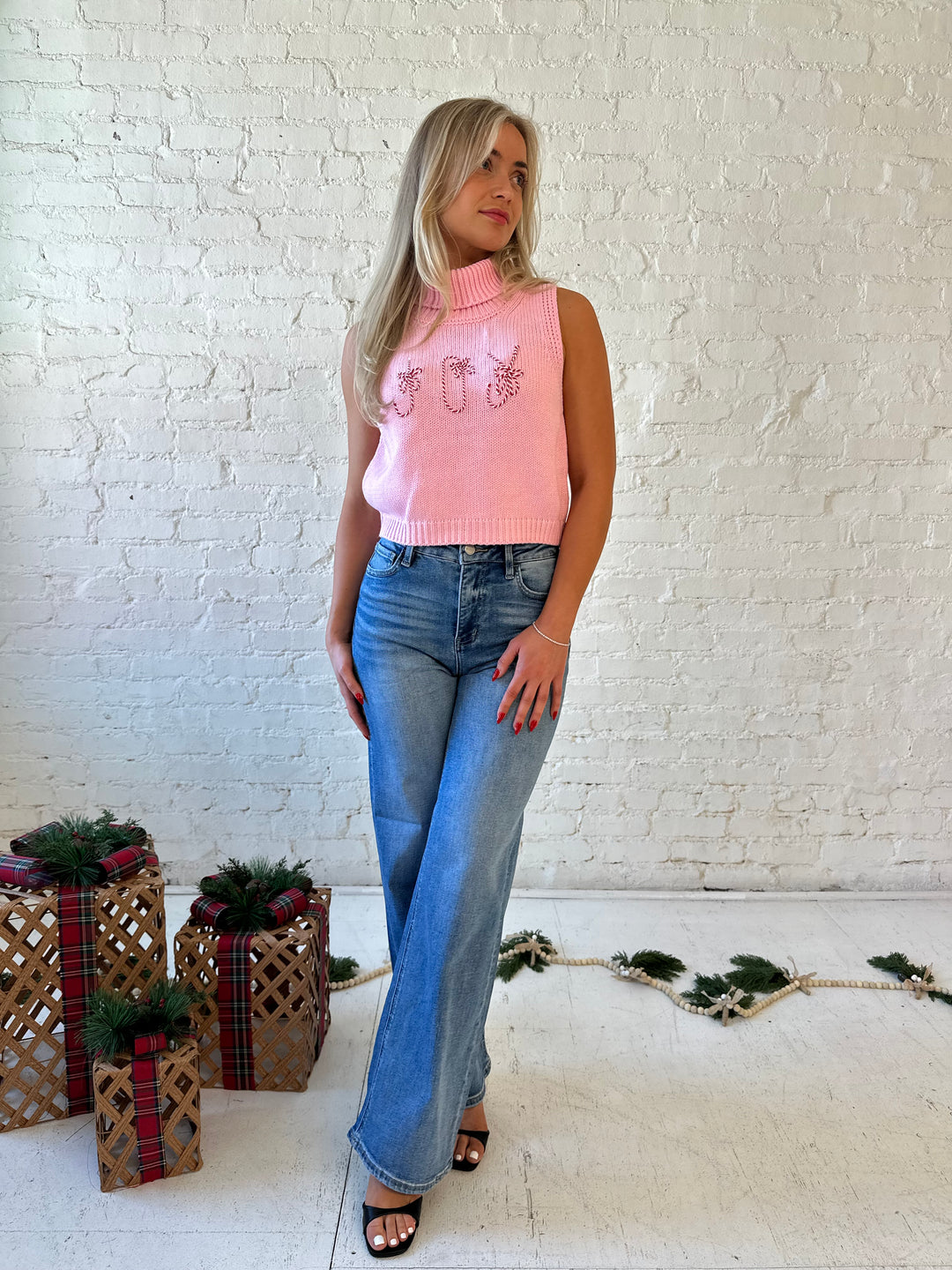 Woman wearing pink sleeveless knit turtleneck top with joy written across the front. 