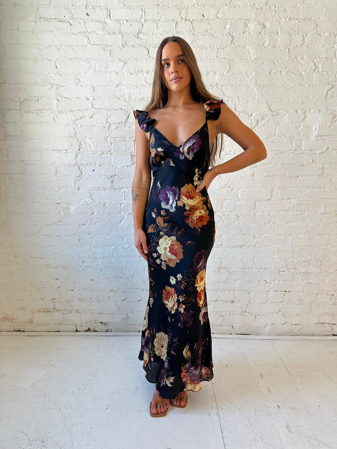 Woman wearing black floral satin maxi dress with v-neckline, ruffle, adjustable spaghetti straps, and open back.