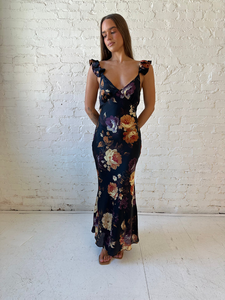 Woman wearing black floral satin maxi dress with v-neckline, ruffle, adjustable spaghetti straps, and open back.