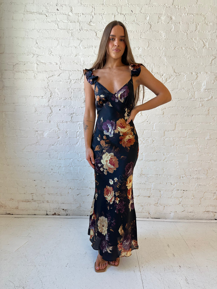 Woman wearing black floral satin maxi dress with v-neckline, ruffle, adjustable spaghetti straps, and open back.