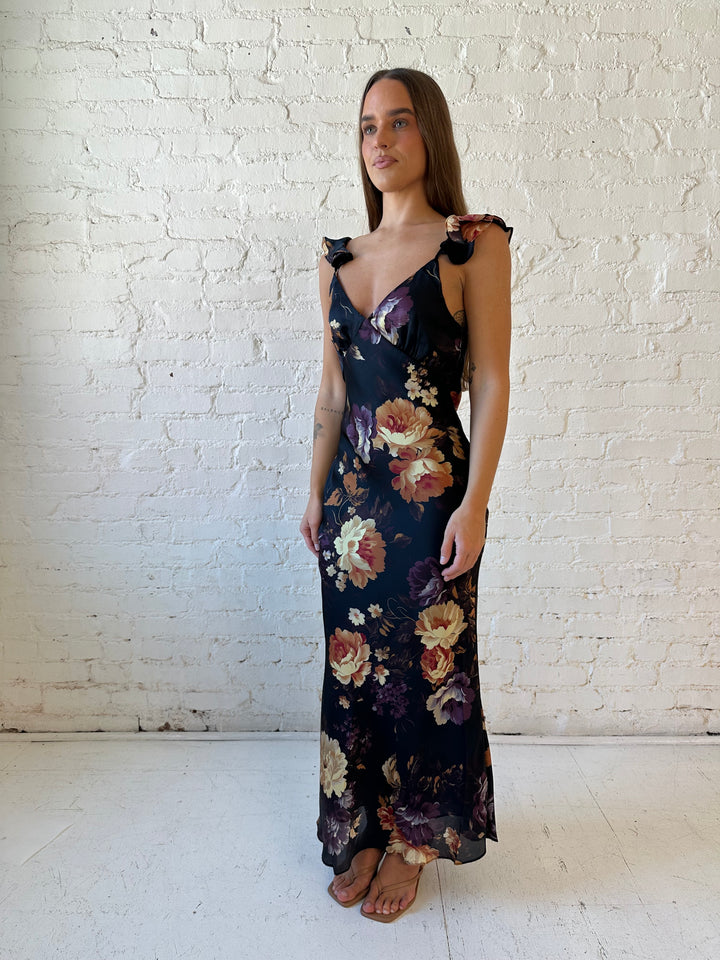 Woman wearing black floral satin maxi dress with v-neckline, ruffle, adjustable spaghetti straps, and open back.