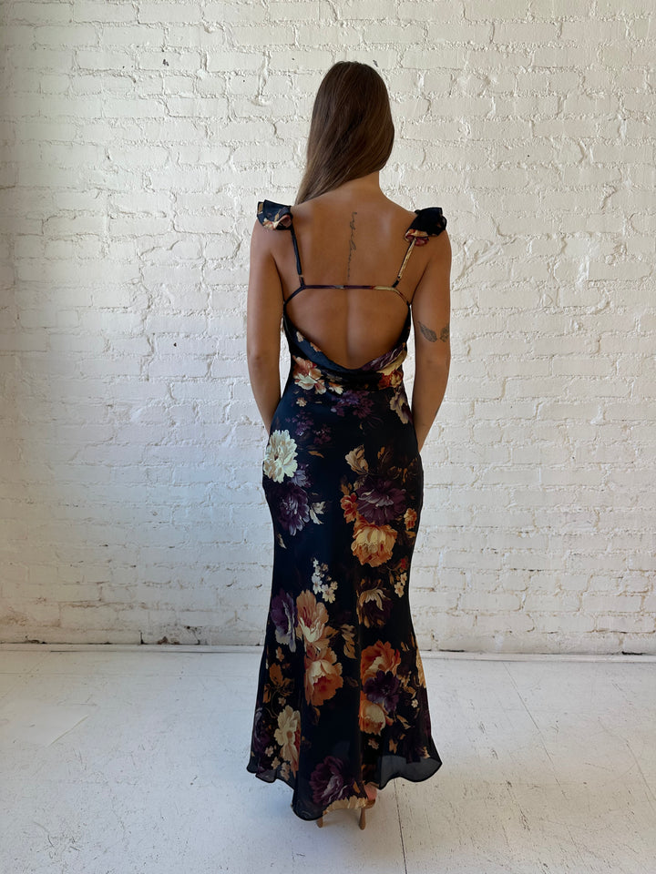 Woman wearing black floral satin maxi dress with v-neckline, ruffle, adjustable spaghetti straps, and open back.