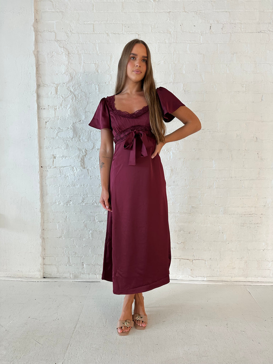 Woman wearing burgundy satin midi dress with lace trim, and self tie detail.