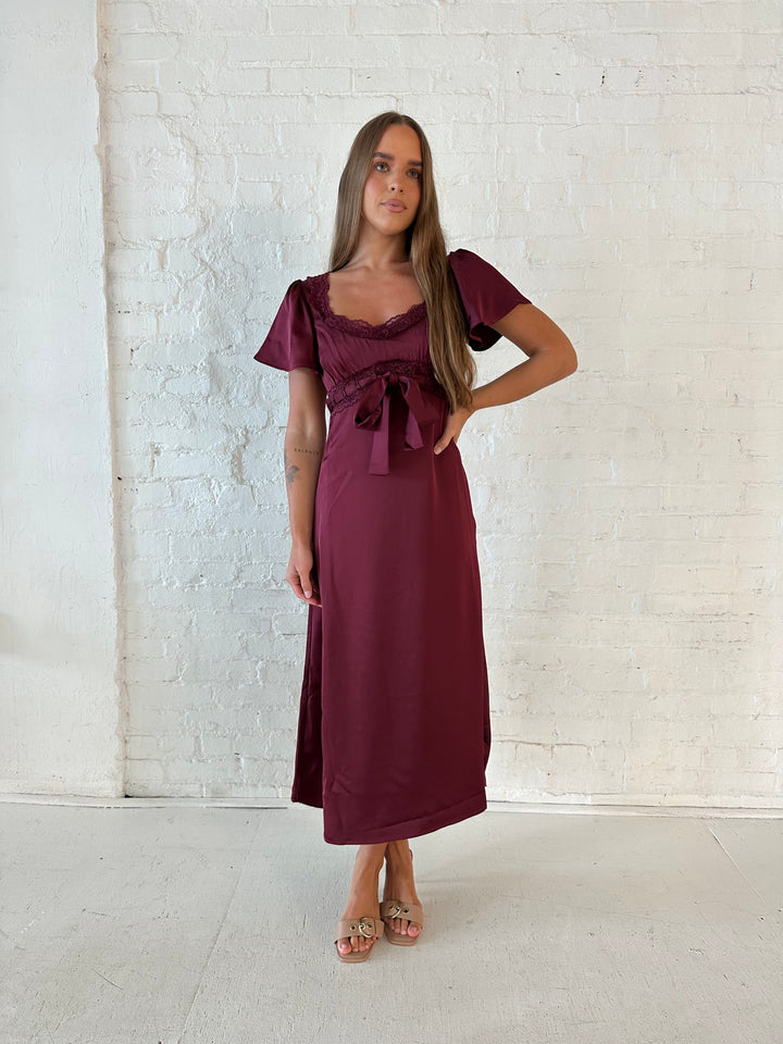 Woman wearing burgundy satin midi dress with lace trim, and self tie detail.
