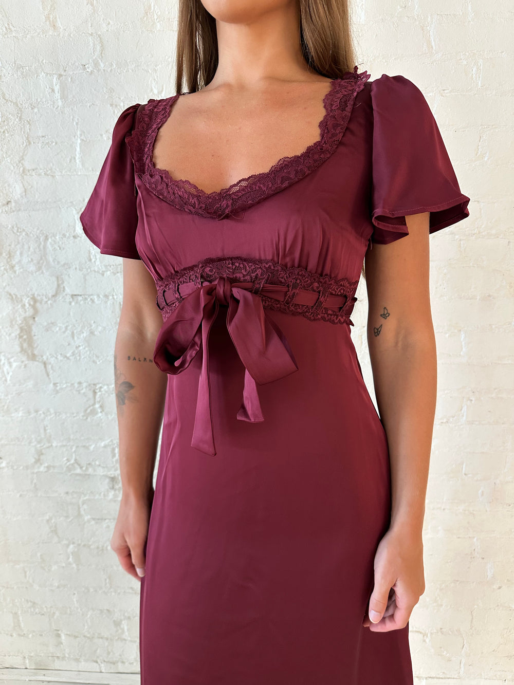 Woman wearing burgundy satin midi dress with lace trim, and self tie detail.
