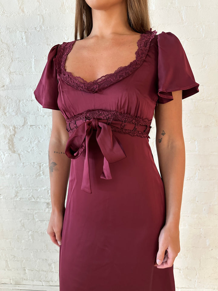 Woman wearing burgundy satin midi dress with lace trim, and self tie detail.