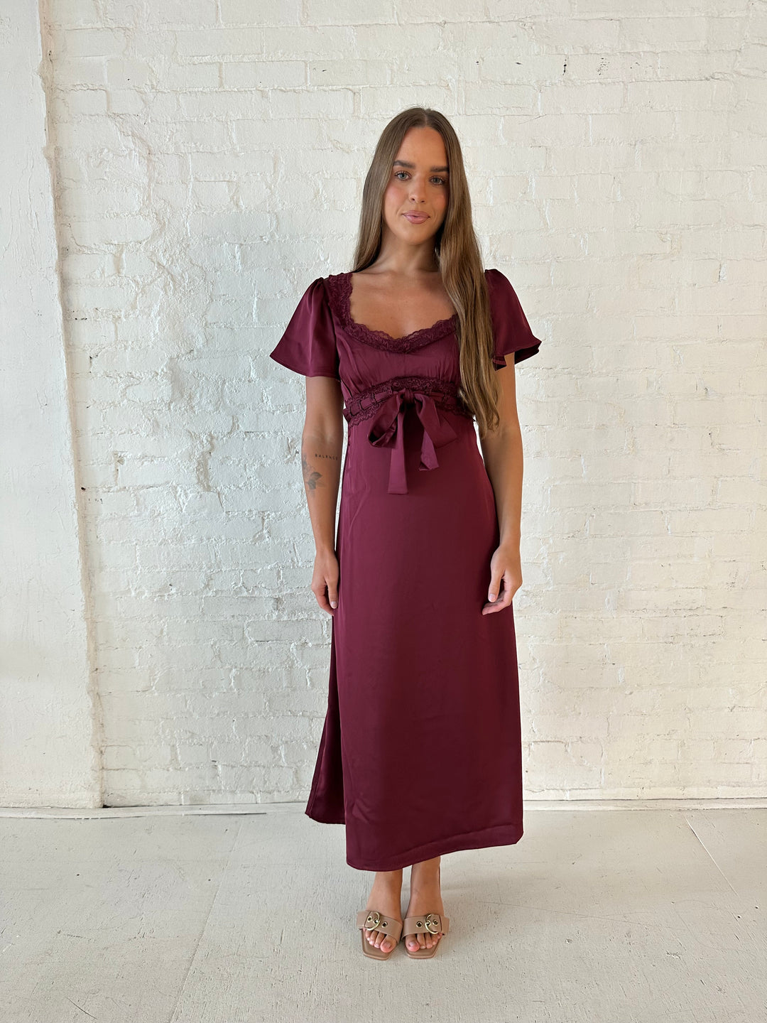 Woman wearing burgundy satin midi dress with lace trim, and self tie detail.