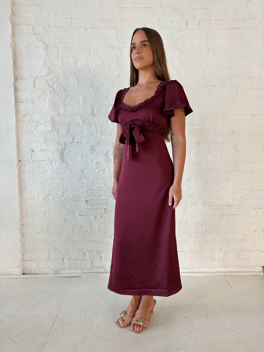 Woman wearing burgundy satin midi dress with lace trim, and self tie detail.