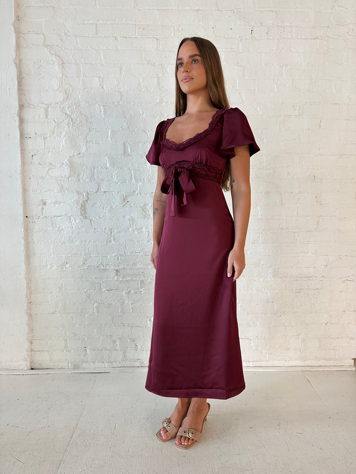 Woman wearing burgundy satin midi dress with lace trim, and self tie detail.