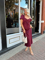 Woman wearing burgundy satin midi dress with puff sleeve, and lace detailing.