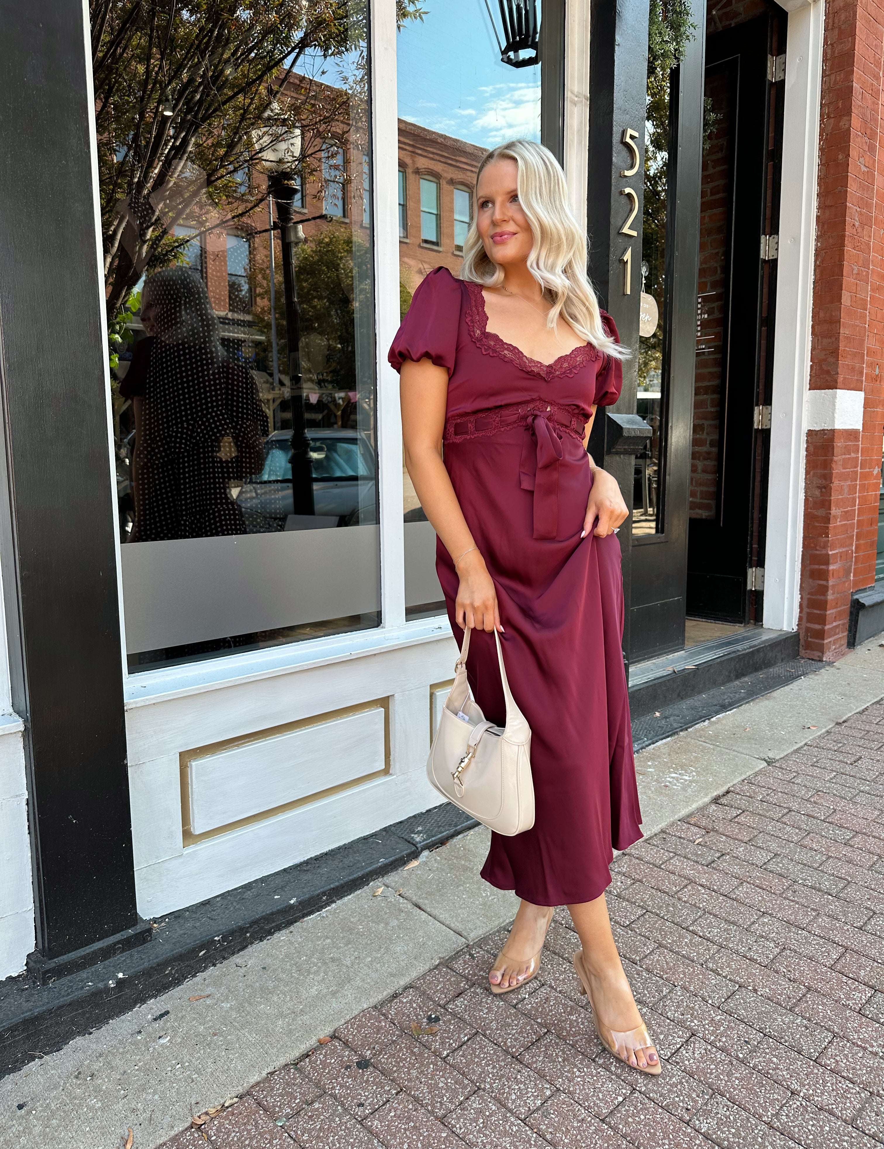 Woman wearing burgundy satin midi dress with puff sleeve, and lace detailing.