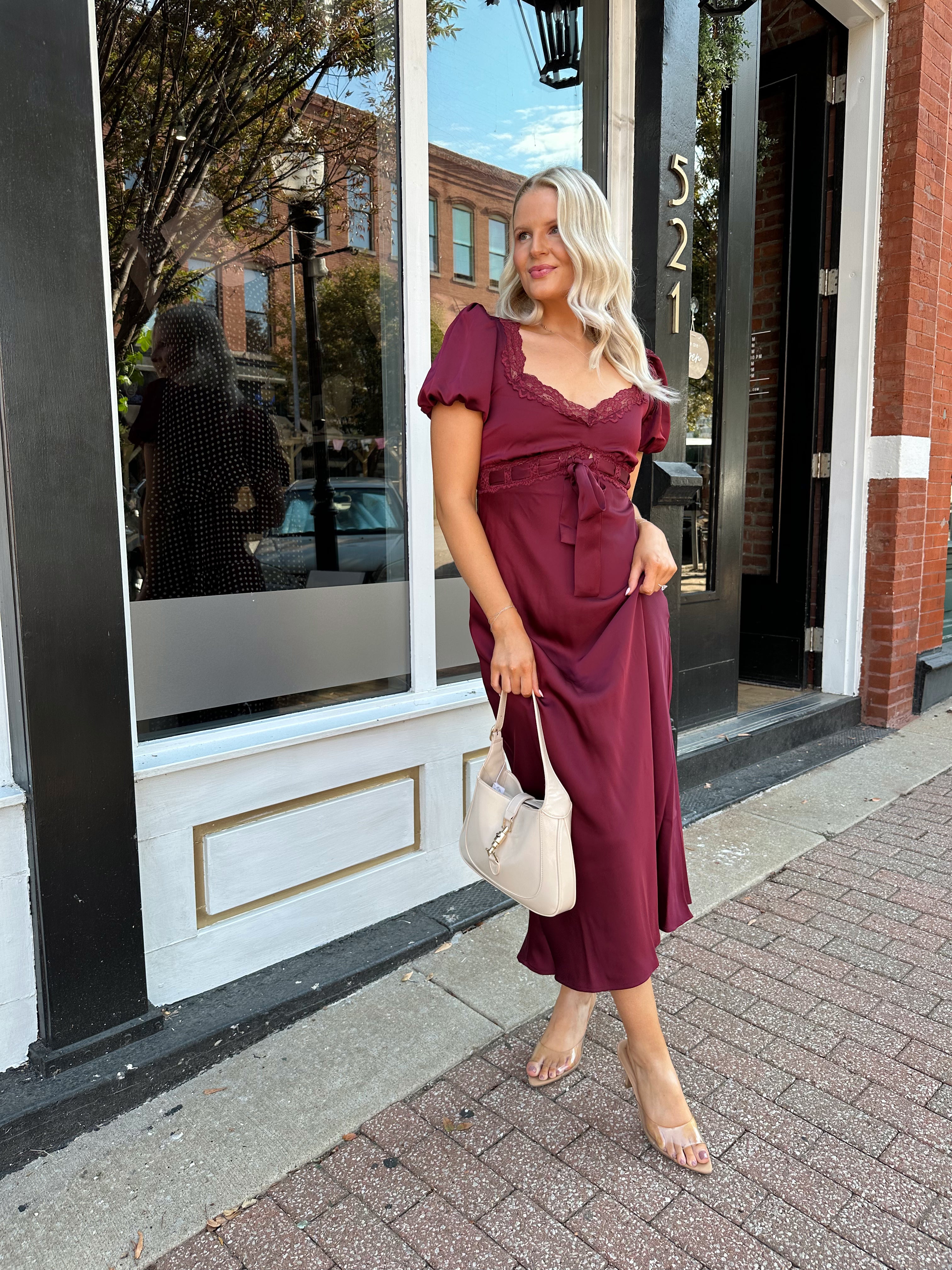 Woman wearing burgundy satin midi dress with puff sleeve, and lace detailing.