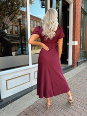 Woman wearing burgundy satin midi dress with puff sleeve, and lace detailing.