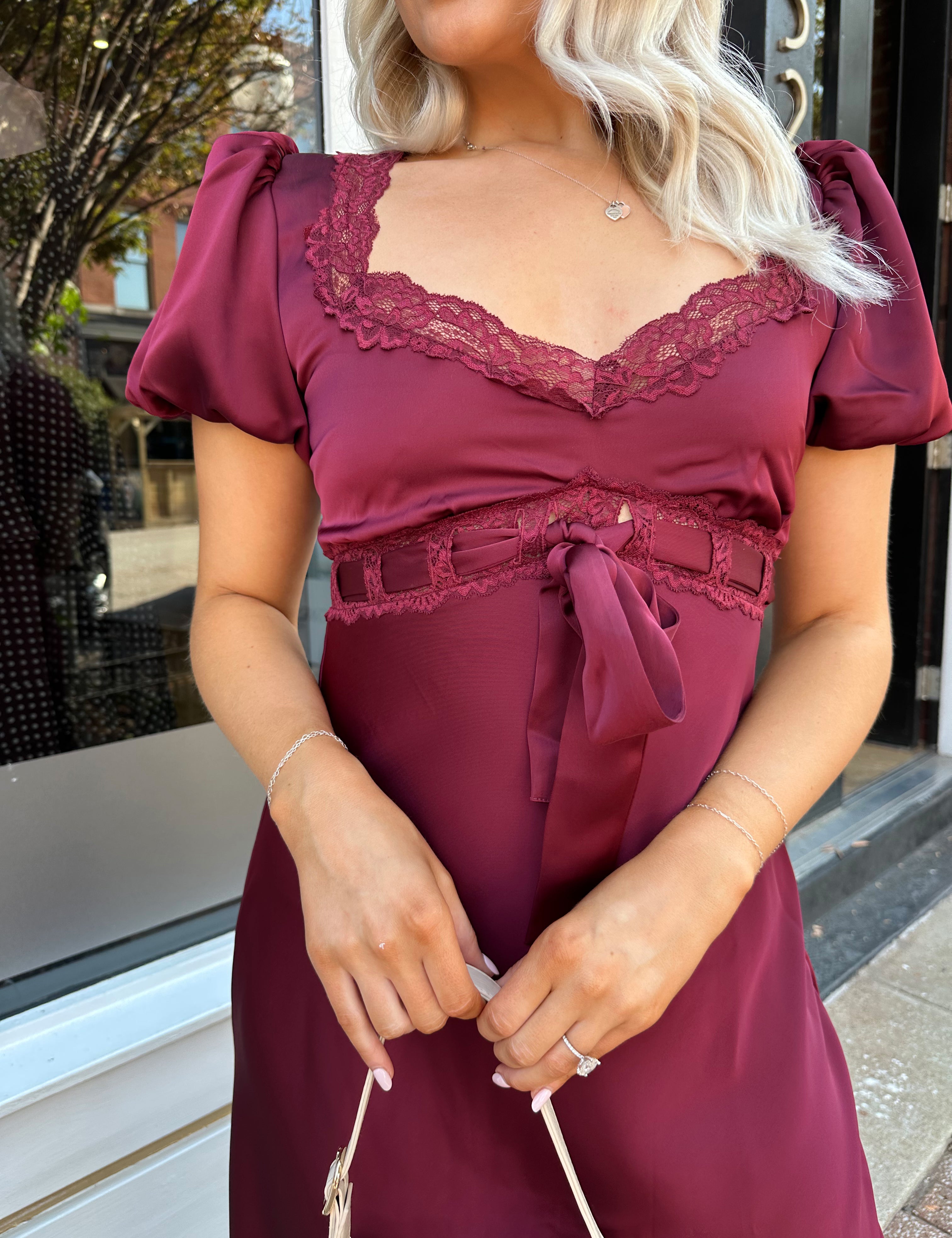Woman wearing burgundy satin midi dress with puff sleeve, and lace detailing.