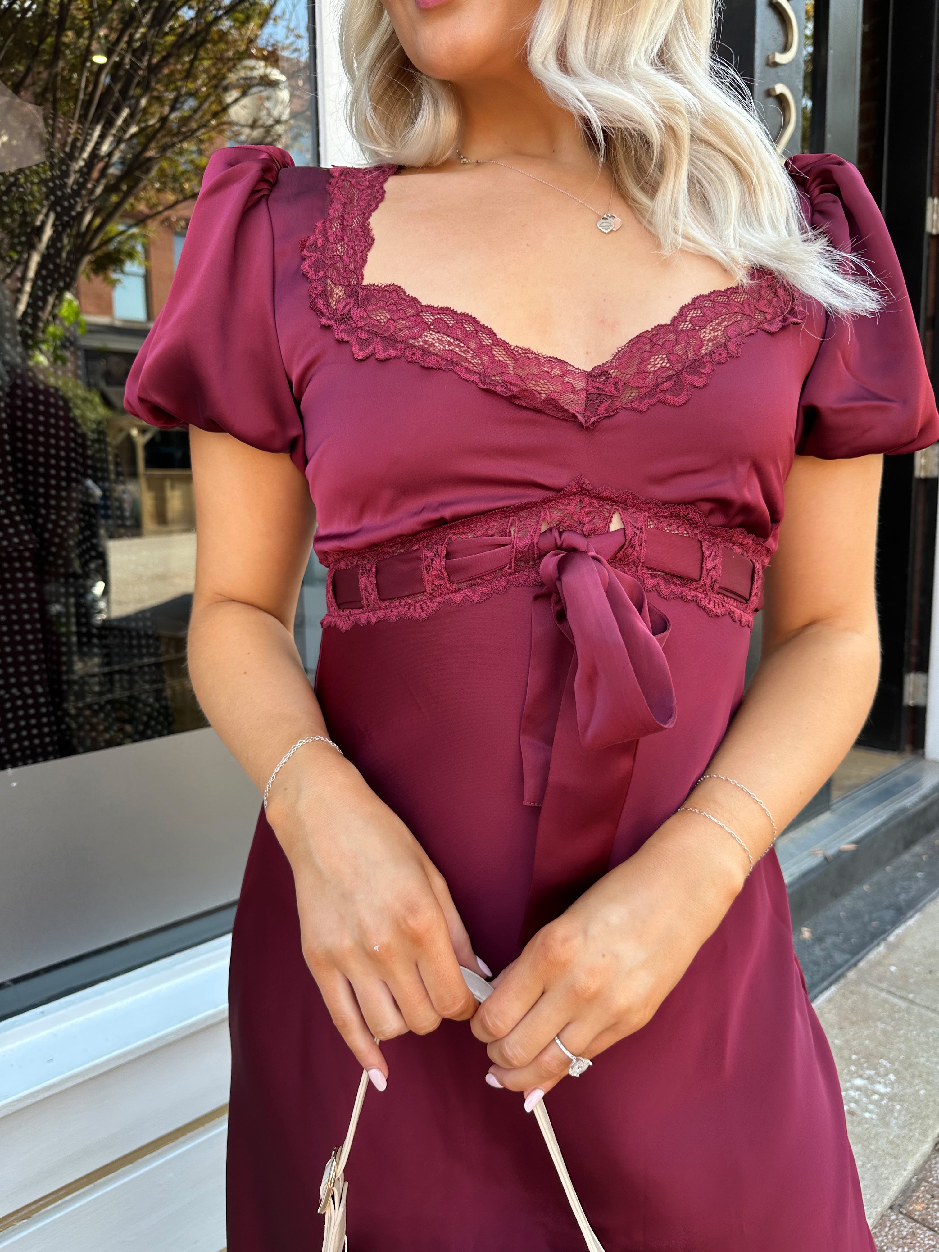 Woman wearing burgundy satin midi dress with puff sleeve, and lace detailing.