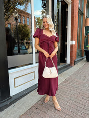 Woman wearing burgundy satin midi dress with puff sleeve, and lace detailing.