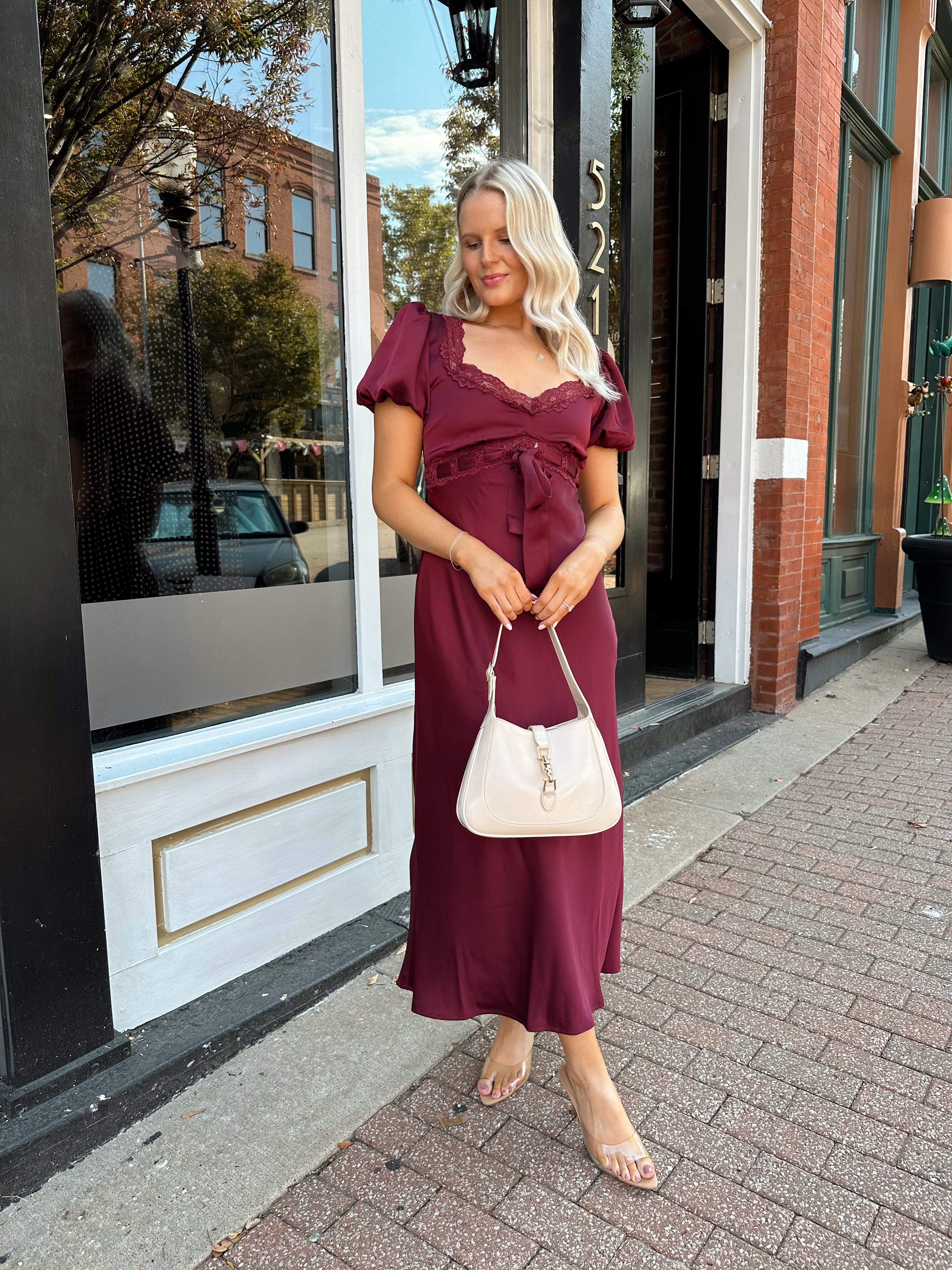 Woman wearing burgundy satin midi dress with puff sleeve, and lace detailing.