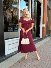 Woman wearing burgundy satin midi dress with puff sleeve, and lace detailing.