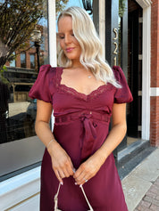 Woman wearing burgundy satin midi dress with puff sleeve, and lace detailing.