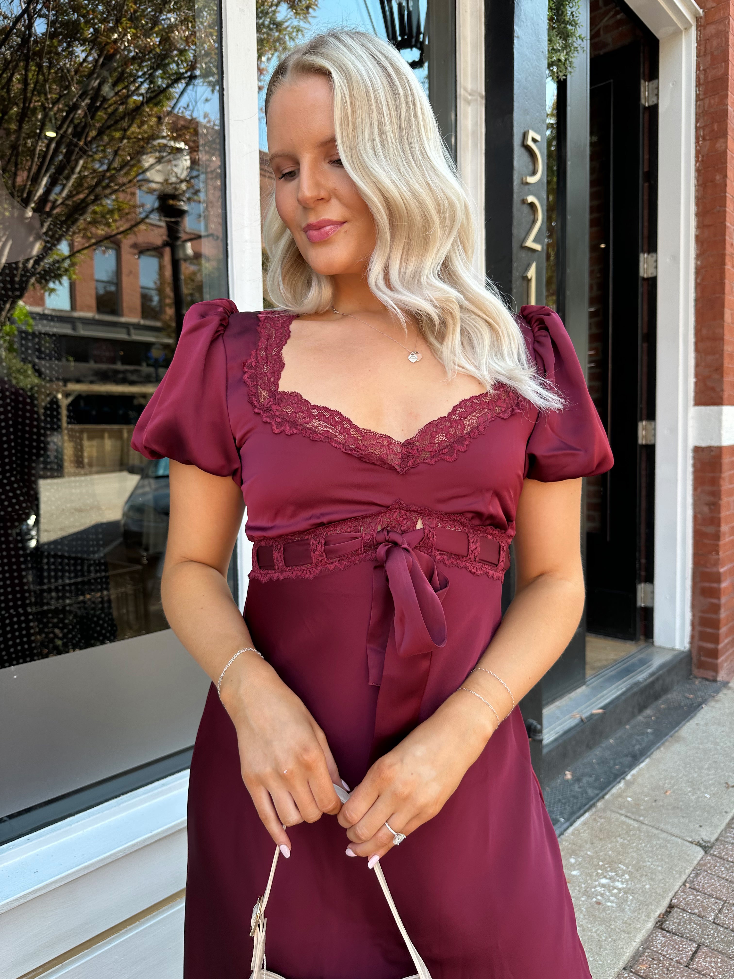 Woman wearing burgundy satin midi dress with puff sleeve, and lace detailing.